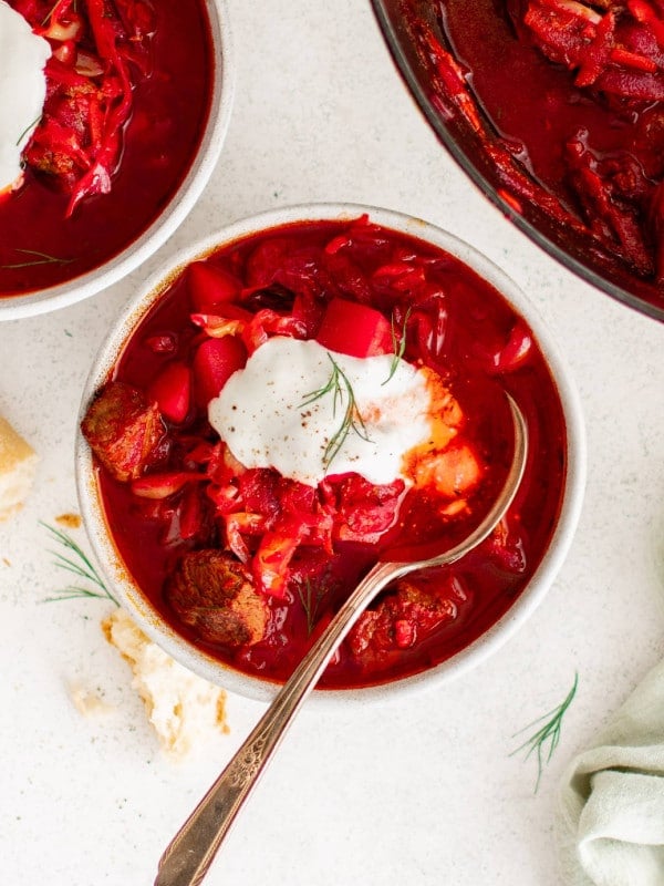 two white bowls filled with borscht soup topped with sour cream set next to a large pot filled with homemade borscht soup.
