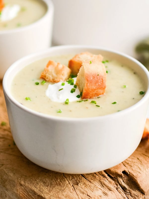White soup bowl on a rustic wood board filled with homemade creamy potato leek soup and garnished with sour cream, homemade croutons, and freshly chopped chives.