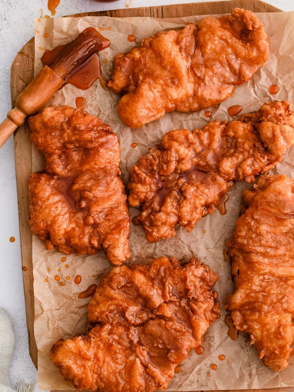 A rustic wooden cutting board piled high with golden-brown fried chicken, each piece generously coated in a glossy hot honey glaze. The sauce glistens under soft lighting, accentuating the crispy texture of the crust. Small drips of honey pool on the board, and a few sprigs of fresh parsley add a pop of color. A small bowl of extra hot honey sits nearby for dipping or drizzling.