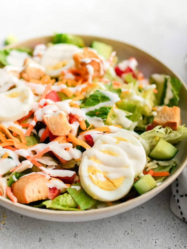 A big, round serving bowl brimming with a colorful house salad, perfectly layered with fresh lettuce, crisp cucumbers, juicy tomatoes, shredded carrots, Cheddar cheese, sliced hard-boiled eggs, and golden croutons, all finished with a generous drizzle of ranch dressing.